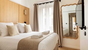 A neatly made bed with white linens and a brown throw, a large mirror on the wall, and a view of the hallway through a window with curtains. - Maison Legendre - Luxury Townhouse with 5BDR & AC in Batignolles (Paris)