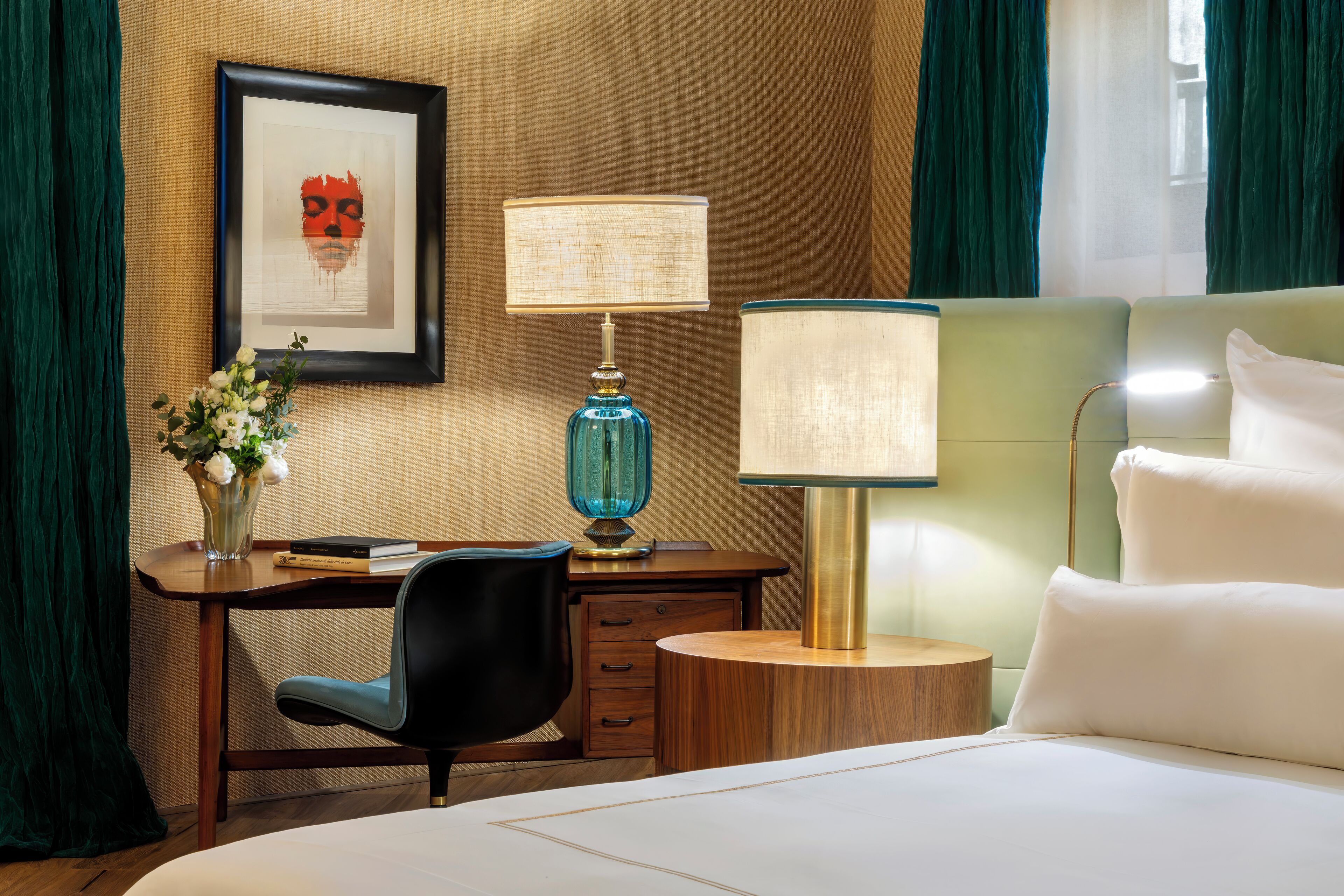 Habitación de hotel con una cama, un escritorio con lámpara, una silla, un florero con flores y un cuadro en la pared.