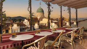 Una terraza al aire libre con mesas redondas y sillas de mimbre, con vistas a un edificio histórico de cúpula verde.