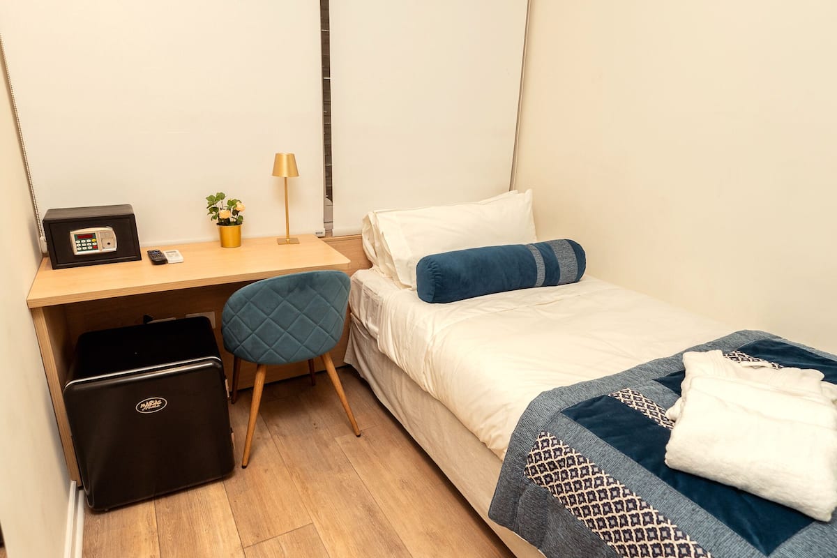 Habitación de hotel compacta con cama, escritorio, silla, una caja de seguridad pequeña y una planta en maceta.