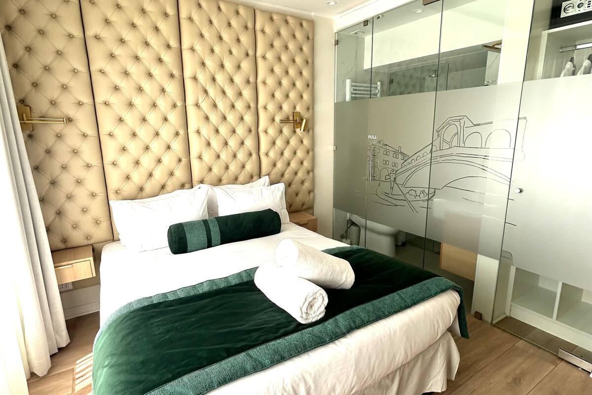 Un dormitorio moderno con un cabecero capitonado, una cama con ropa de cama verde y blanca, y un armario con puertas de cristal que muestra un mural de un paisaje urbano.
