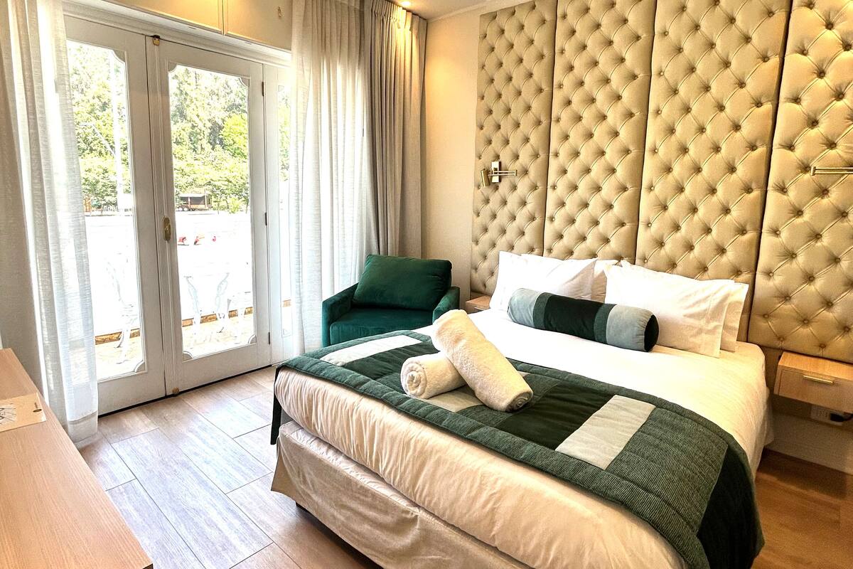 Habitación de hotel con una cama grande, un sillón verde, un escritorio de madera y vistas a través de puertas de cristal.