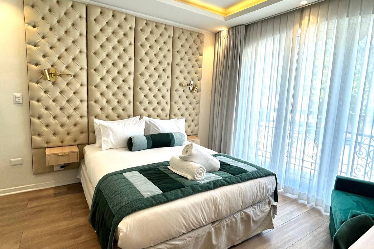 Una habitación de hotel moderna con una cama grande, cabecera con diseño acolchado, mesita de noche y vistas al exterior a través de cortinas traslúcidas.