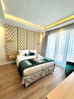 Une chambre d’hôtel moderne avec un grand lit, une tête de lit à motifs capitonnés, une table de chevet et une vue sur l’extérieur à travers des rideaux transparents.