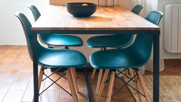 Un espace repas avec une table en bois, des chaises de couleur turquoise, un luminaire suspendu et une vue sur le paysage urbain à travers de grandes fenêtres.