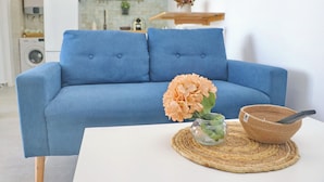 Um sofá azul com duas almofadas, um vaso de vidro com flores e um jogo americano trançado sobre uma mesa branca.