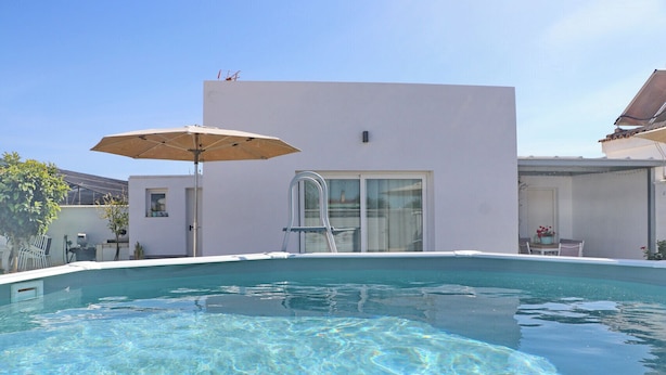 Uma piscina com superfície de água azul límpida, um edifício branco com porta de vidro e uma área de pátio com mesa e cadeiras.