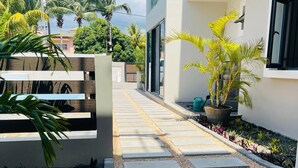 A pathway leading to a modern building with a minimalist design, surrounded by tropical plants and a clear sky. - RABBIT HOLIDAY - 8 mins walk to the beach (Flic en Flac)
