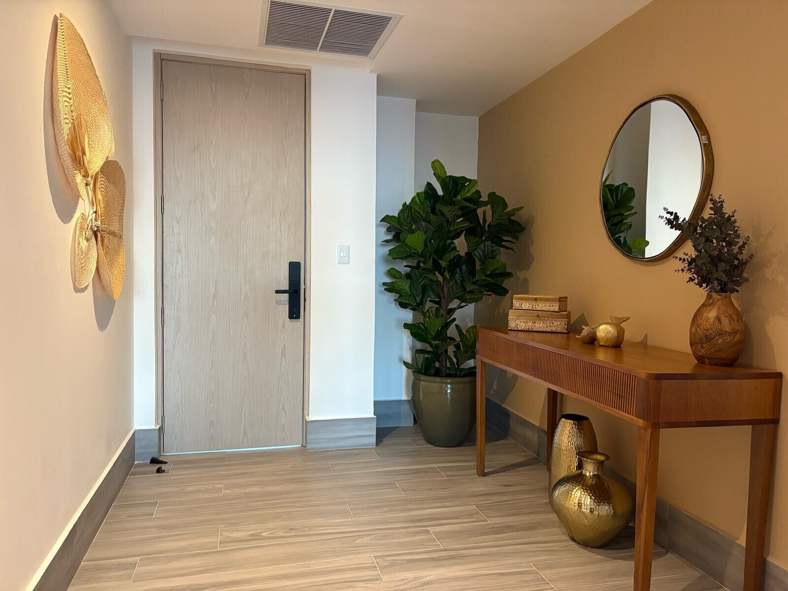 A hallway with a wooden door, a large potted plant, a console table with decorative items, and a wall-mounted mirror.