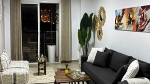 Een moderne woonkamer met een zwarte bank, een houten salontafel en een witte fauteuil. Er is een groot raam met gordijnen, een plant en een schilderij aan de muur.