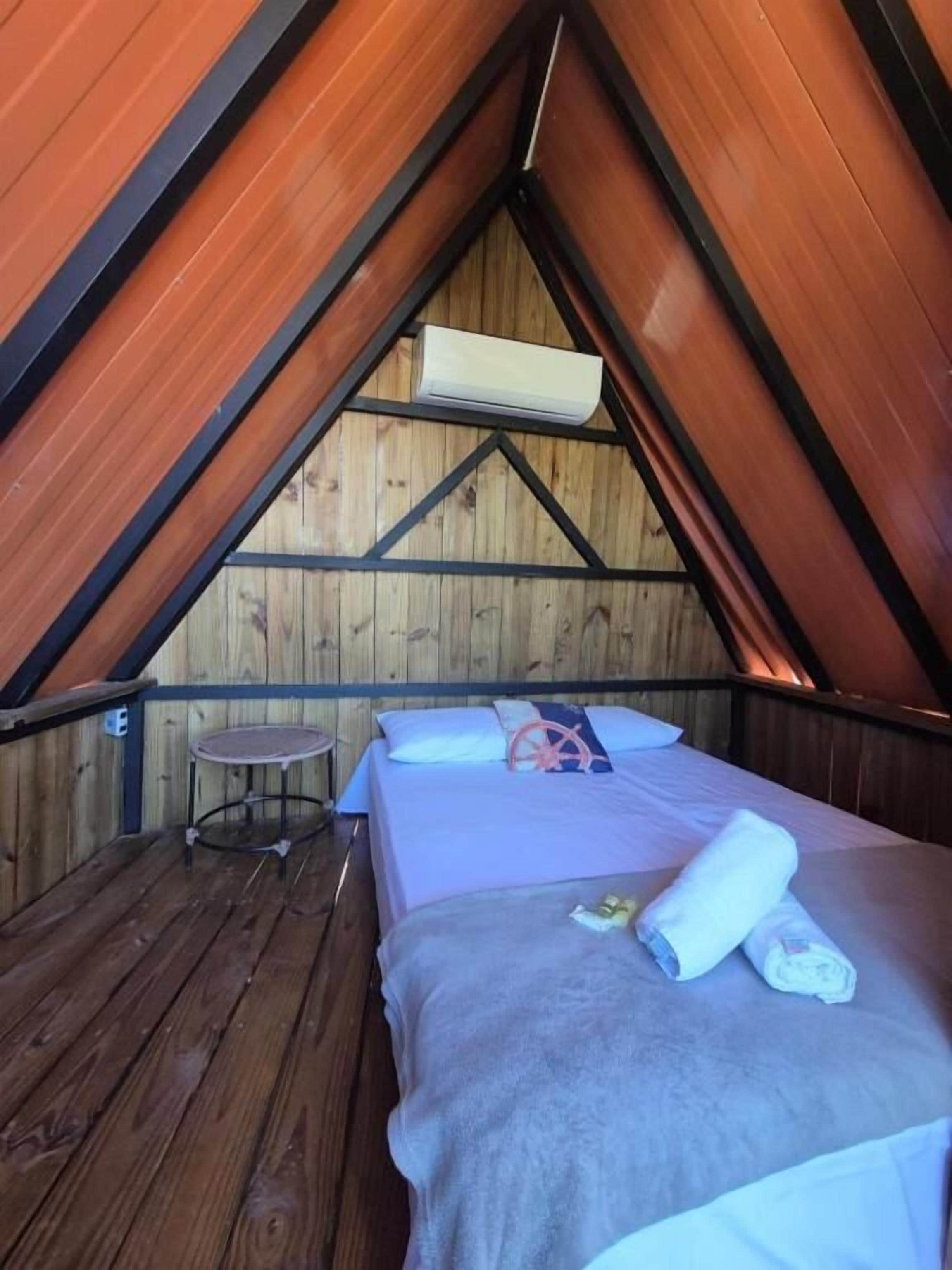 Un dormitorio cálido en el ático, con techo inclinado de madera, una cama individual con almohadas y una toalla enrollada, una pequeña mesa redonda y un equipo de aire acondicionado.