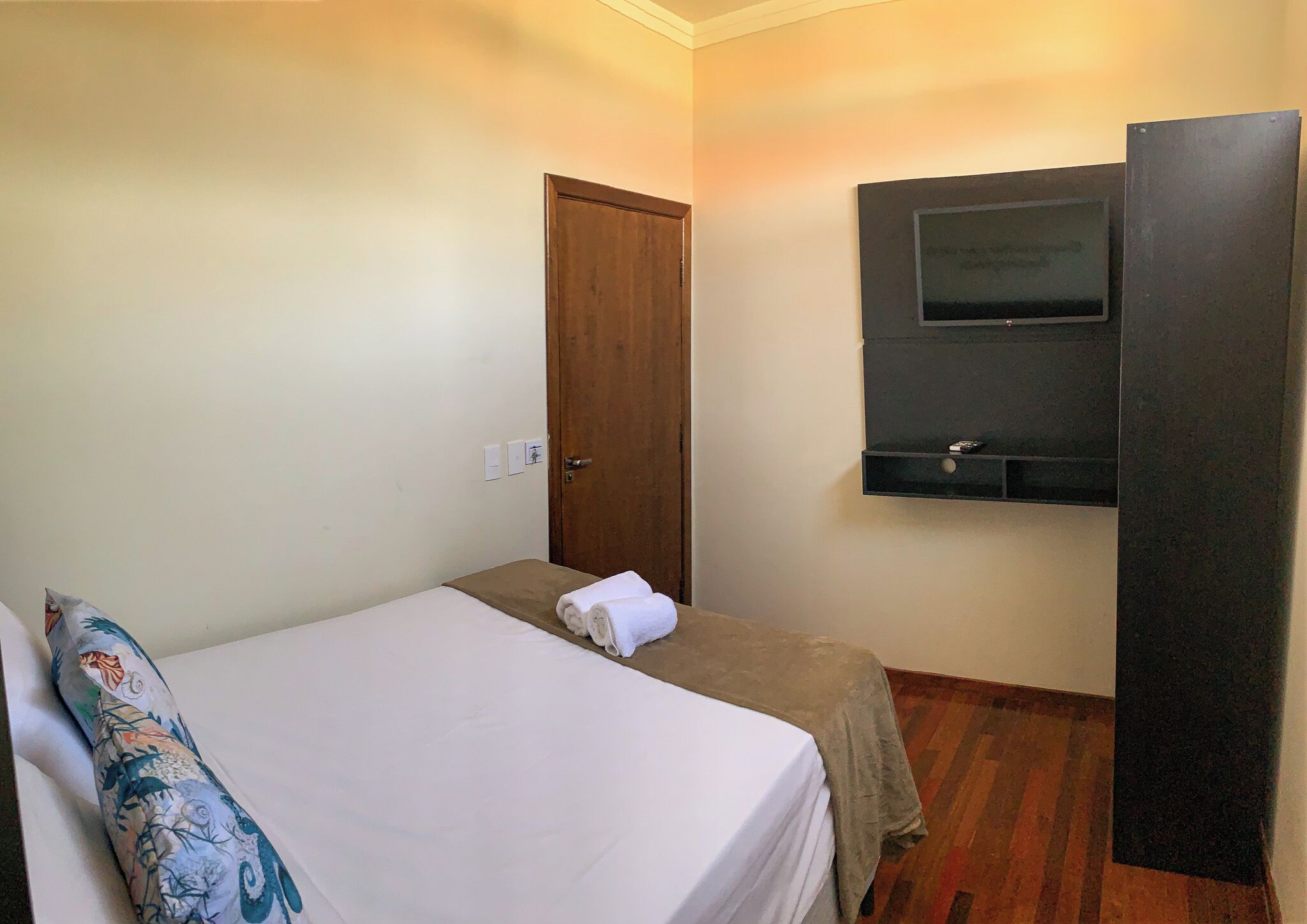 Camera d'albergo con un letto singolo, una porta in legno, una TV fissata al muro e un piccolo ripiano sottostante.