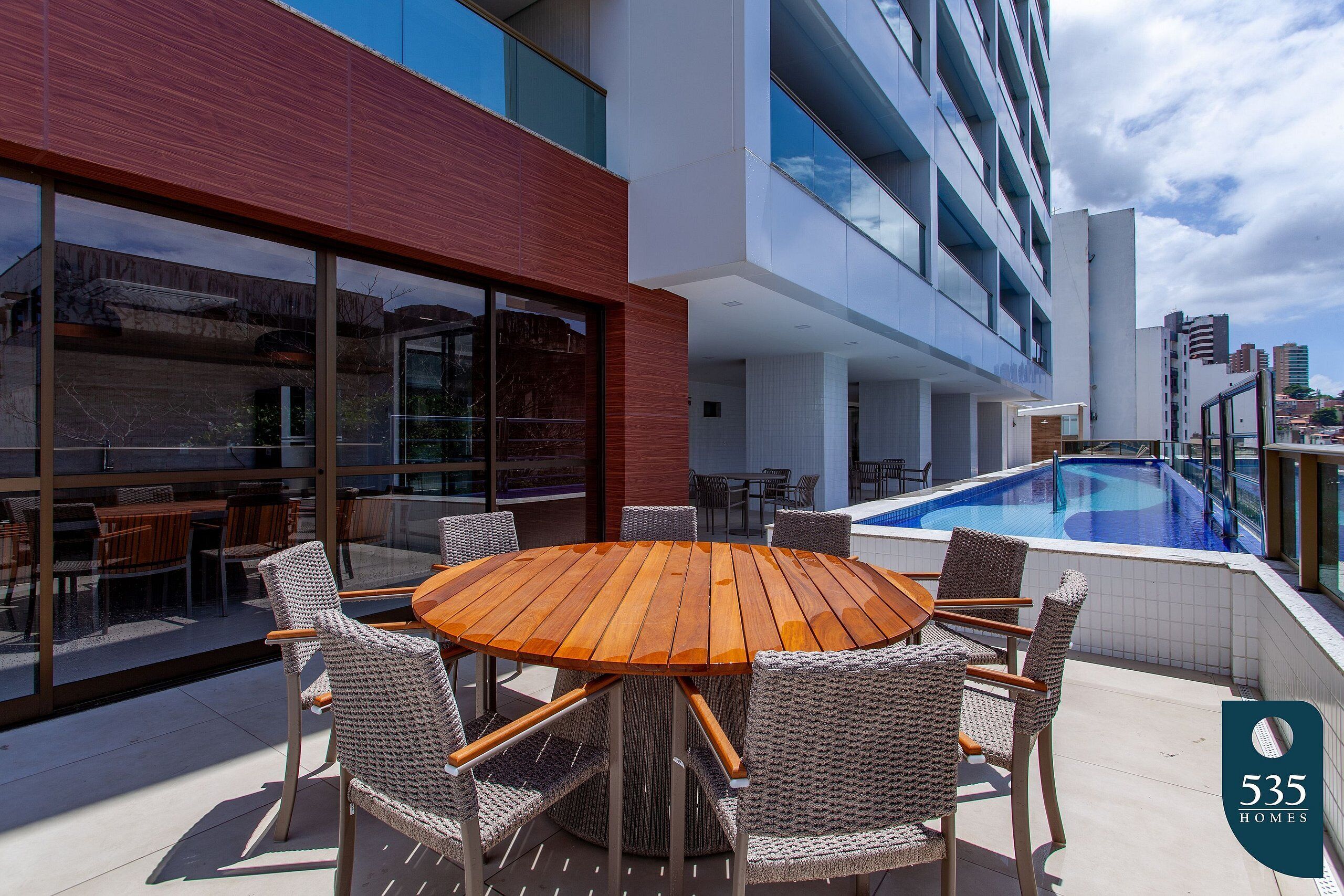 A modern outdoor patio with a wooden table and wicker chairs, a swimming pool, and a glass-enclosed dining area.