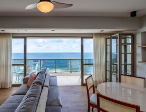 Un salon moderne doté d’un grand canapé, d’une table à manger et offrant une vue sur l’océan grâce à de grandes fenêtres du sol au plafond.