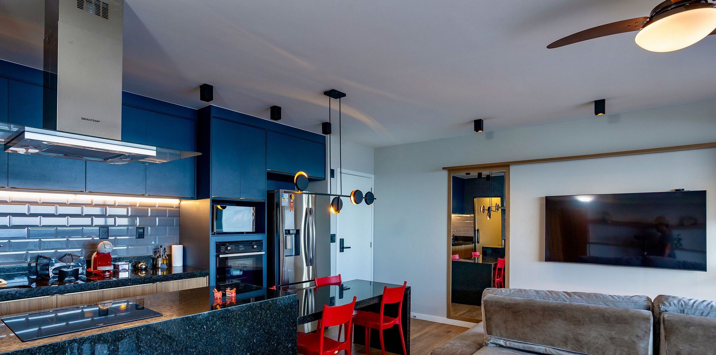 Cocina moderna con refrigerador de acero inoxidable, microondas y horno empotrado. Hay un comedor con mesa de vidrio y sillas rojas. Un televisor de pantalla plana grande está montado en la pared.