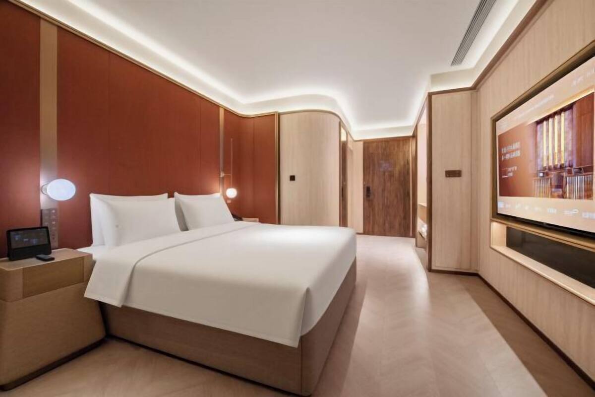 Ein modernes Hotelzimmer mit einem großen Bett, einem Nachttisch mit Lampe, einem an der Wand montierten Fernseher und einer Holzvertäfelung.