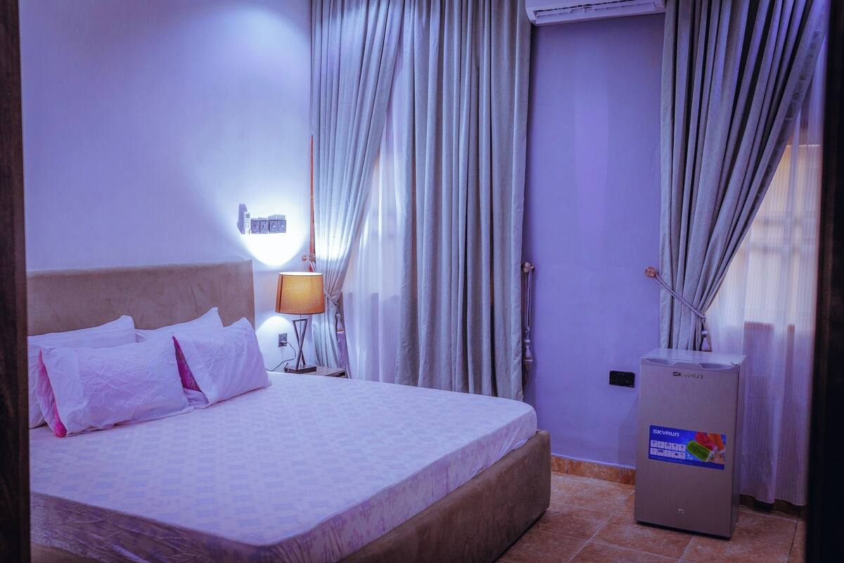 A hotel room with a bed, a nightstand, a lamp, and an air conditioning unit.