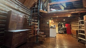 Interior rústico de una cabaña con una escalera de madera que sube a un altillo, una zona de cocina con un refrigerador rojo y un aparador de madera.