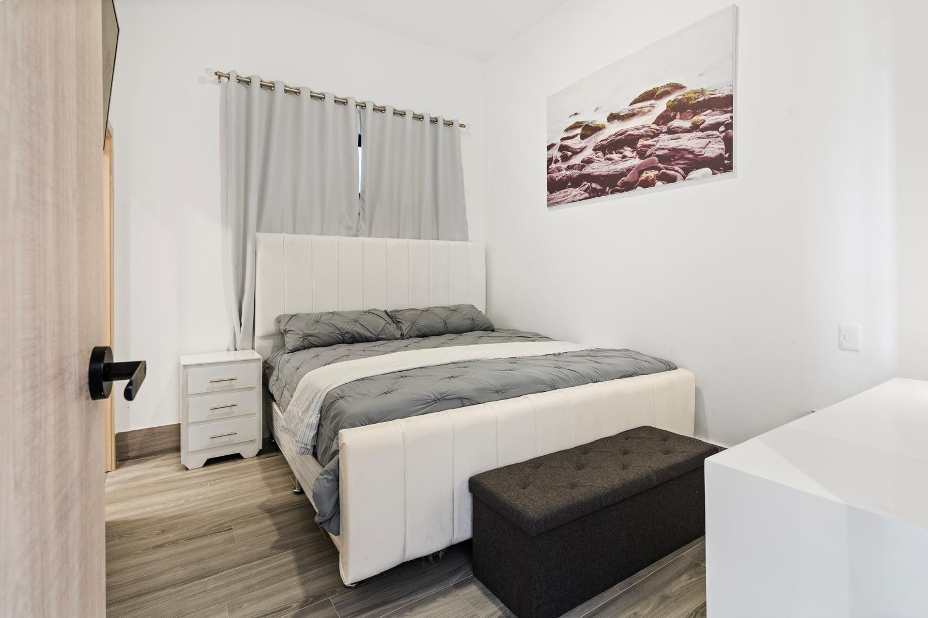 A modern bedroom with a bed, bedside table, bench, and a large window with curtains.
