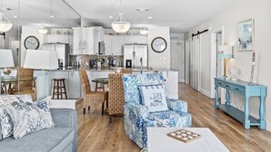 Une cuisine moderne avec des meubles blancs, un coin repas avec des chaises en osier et un salon avec un fauteuil bleu et un canapé gris.