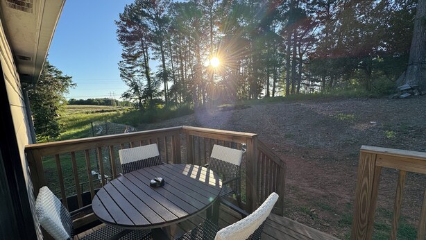 Une terrasse en bois avec une table ronde et quatre chaises, donnant sur une forêt et un chemin de gravier.