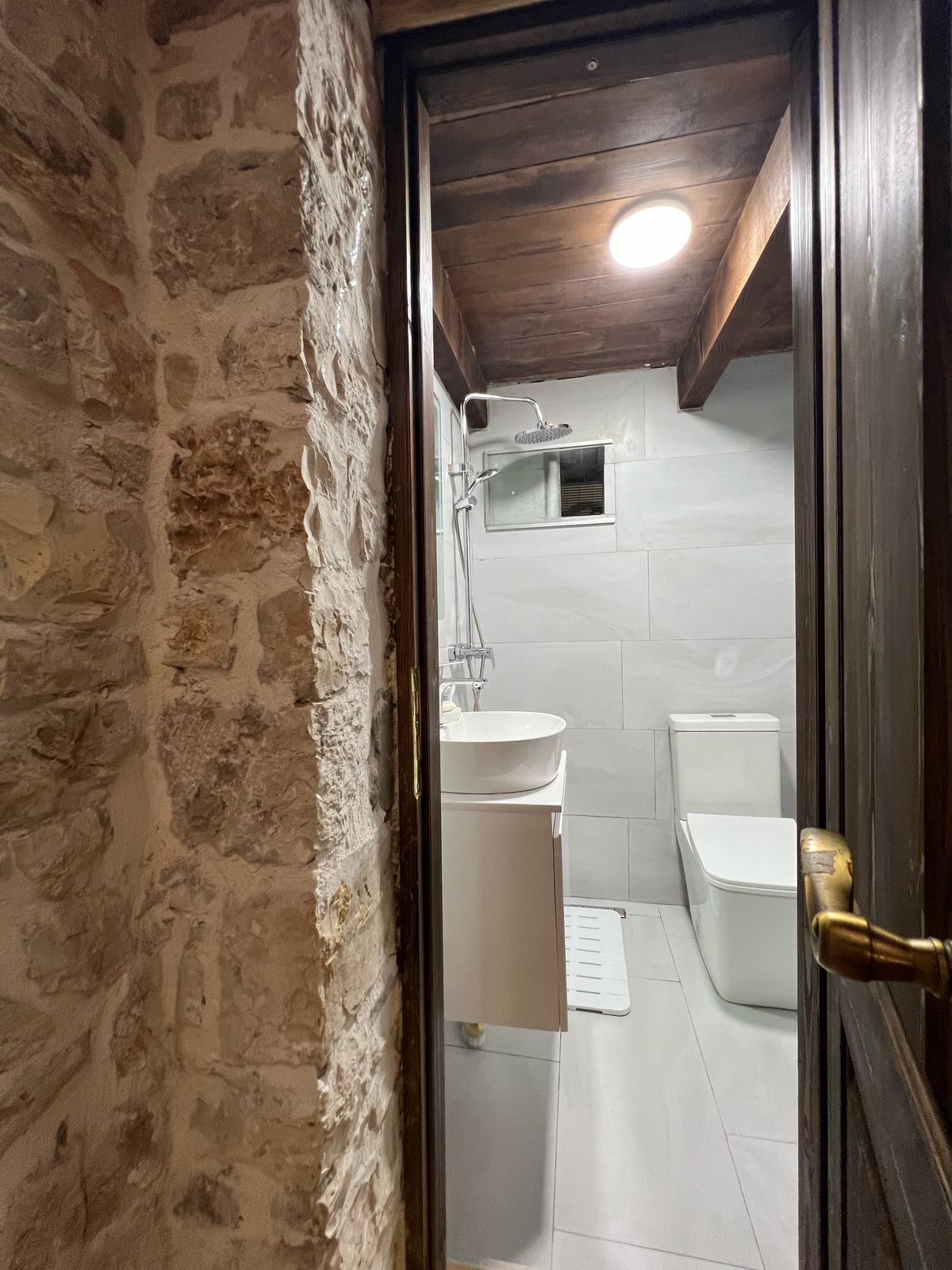 A bathroom with a stone wall, a wooden ceiling, a white sink, a toilet, and a shower.