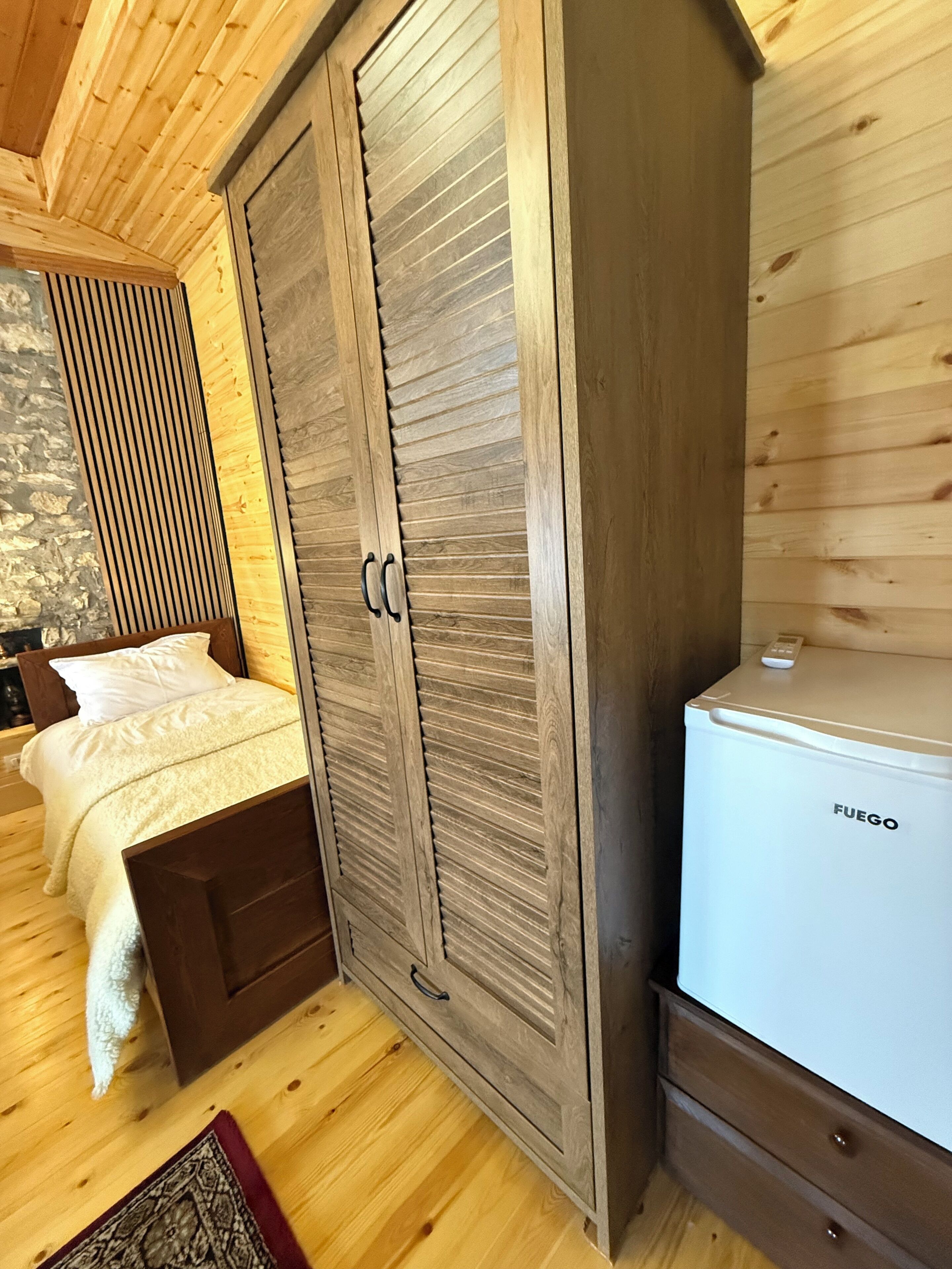 A bedroom with a wooden wardrobe, a bed with white bedding, and a white Fuego refrigerator.