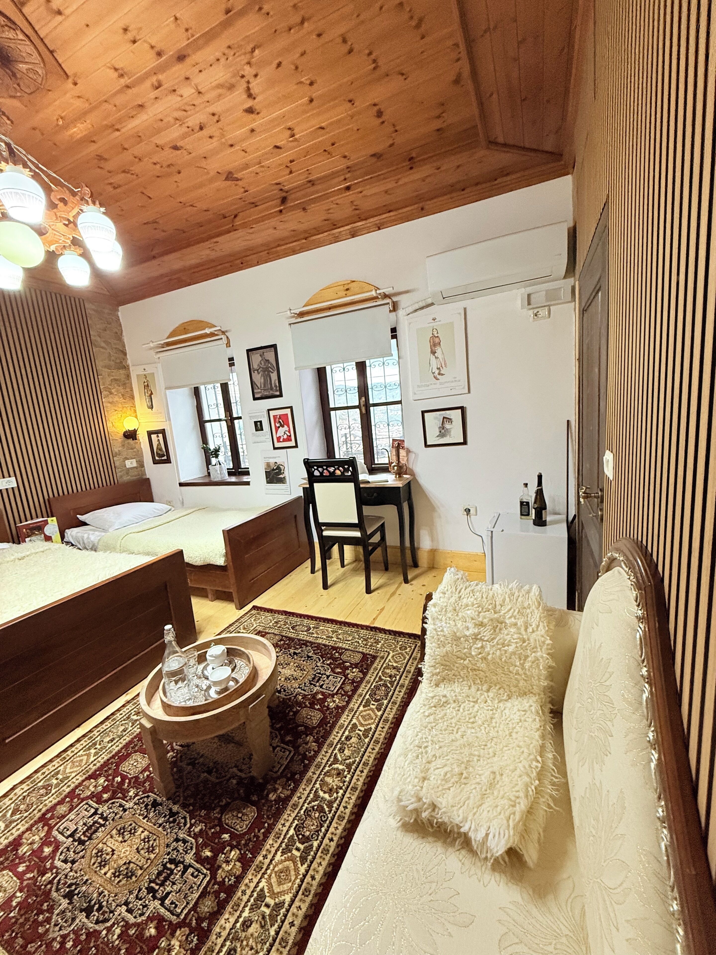 Una habitación acogedora con techo de madera, una cama, un escritorio y un sofá. Hay cuadros enmarcados en la pared y una ventana con vistas.