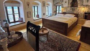 Une chambre avec un grand lit, un plafond en bois et un mur en pierre. On y trouve des fenêtres à arc en plein cintre et un lustre.