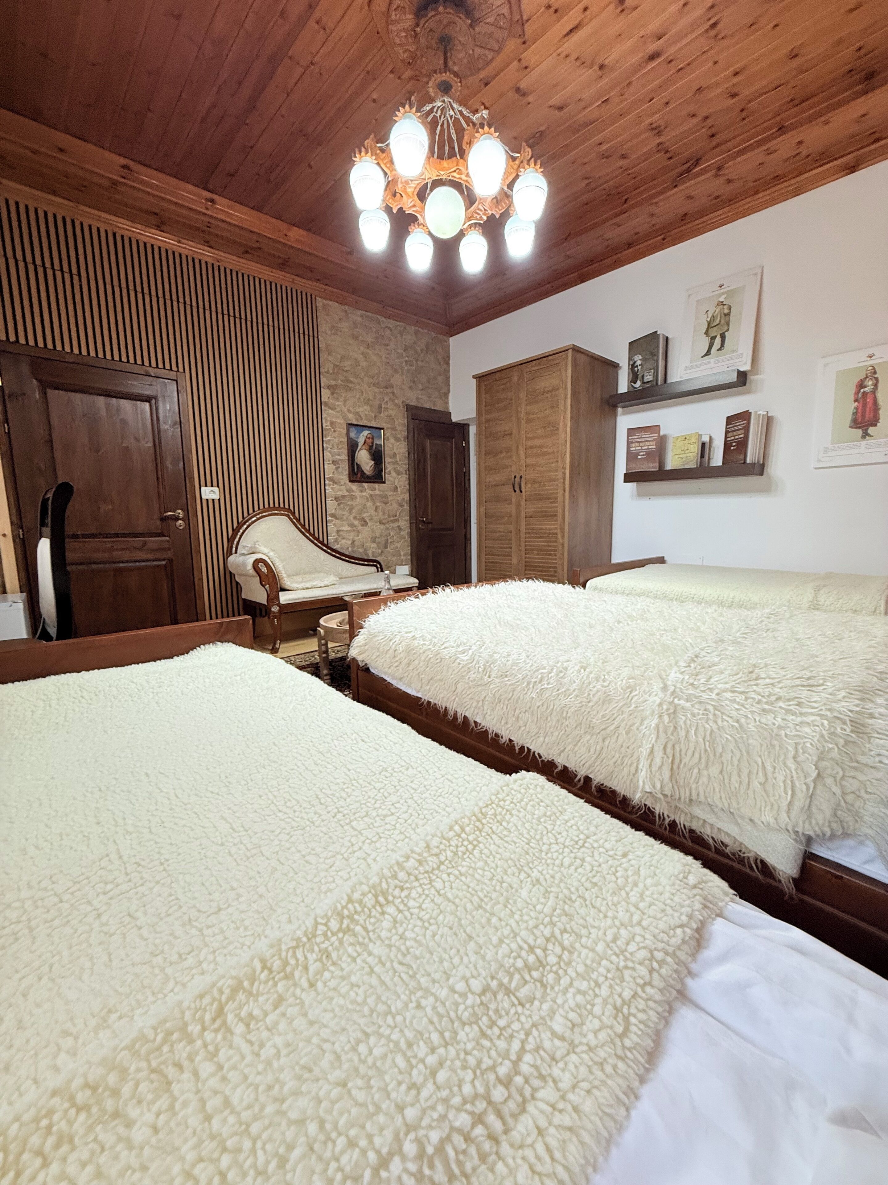 A bedroom with two beds, wooden ceiling, a chandelier, a wardrobe, and a painting on the wall.