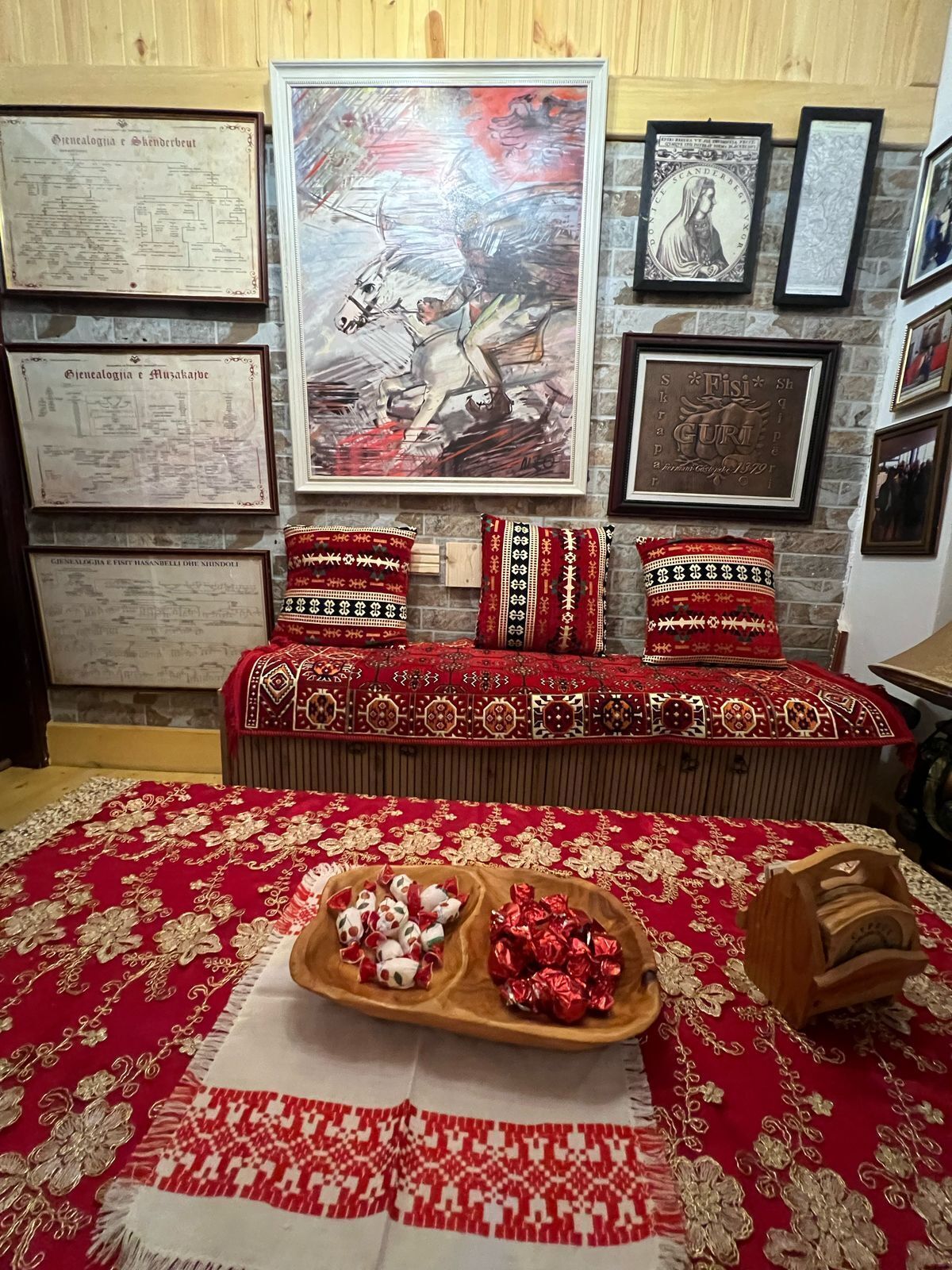 Una habitación con una colcha roja estampada, un banco de madera con cojines, y una pared decorada con documentos enmarcados y un cuadro grande.