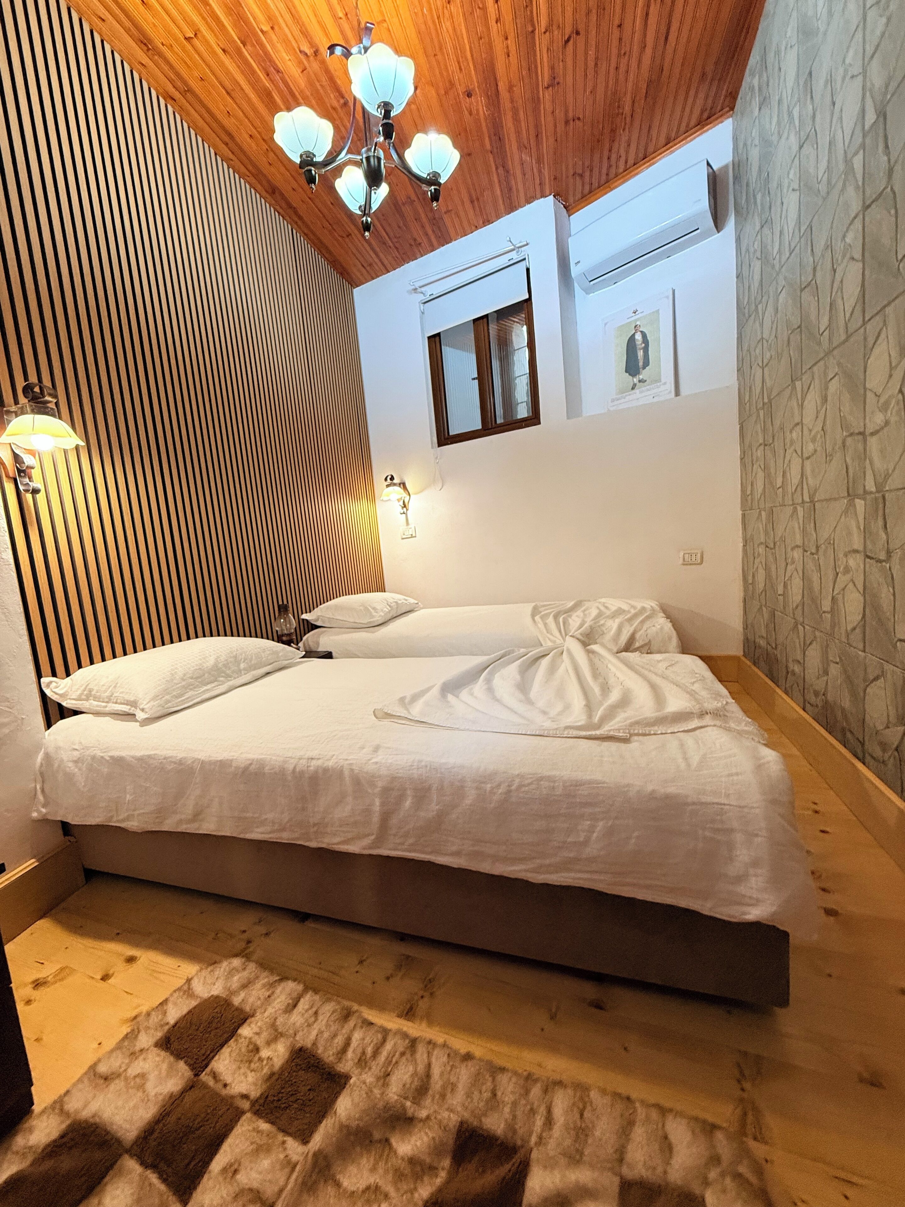Un dormitorio con techo de listones de madera, un candelabro, una cama con ropa blanca, un ventanal pequeño y un aire acondicionado instalado en la pared.
