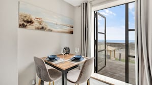 Ein Essbereich mit Tisch für zwei Personen, einem Bild an der Wand und Blick auf den Strand durch ein großes Fenster.