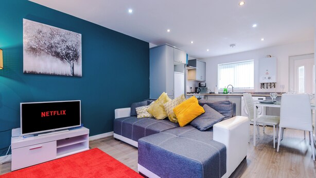 A modern living room with a sectional sofa, a flat-screen TV displaying 'NETFLIX', a red rug, and a painting of a tree on the wall. - 3 Bed-sleeps 5-garden-parking (Liverpool)