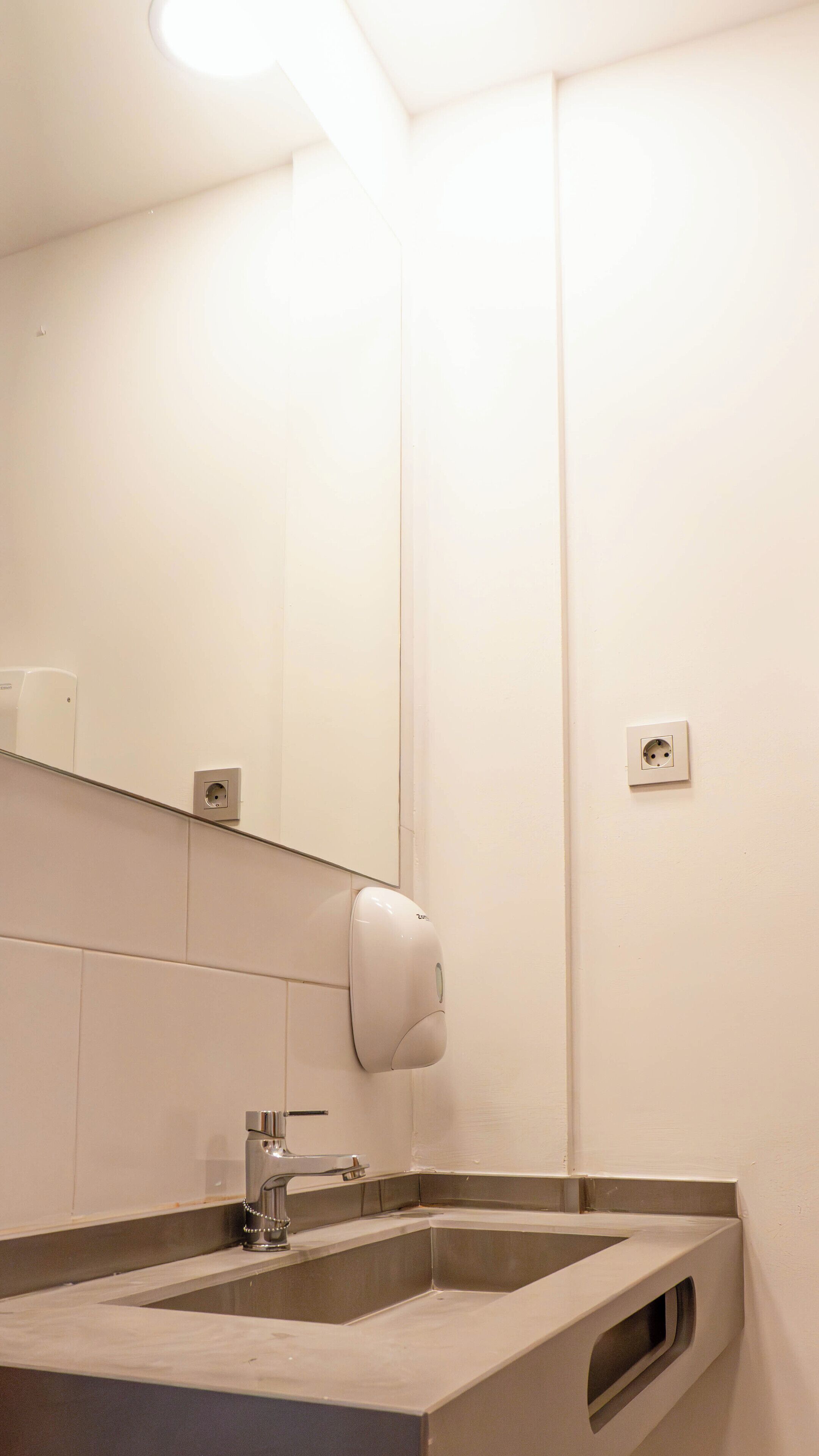 Baño con lavabo, espejo, dispensador de jabón y toma de corriente.