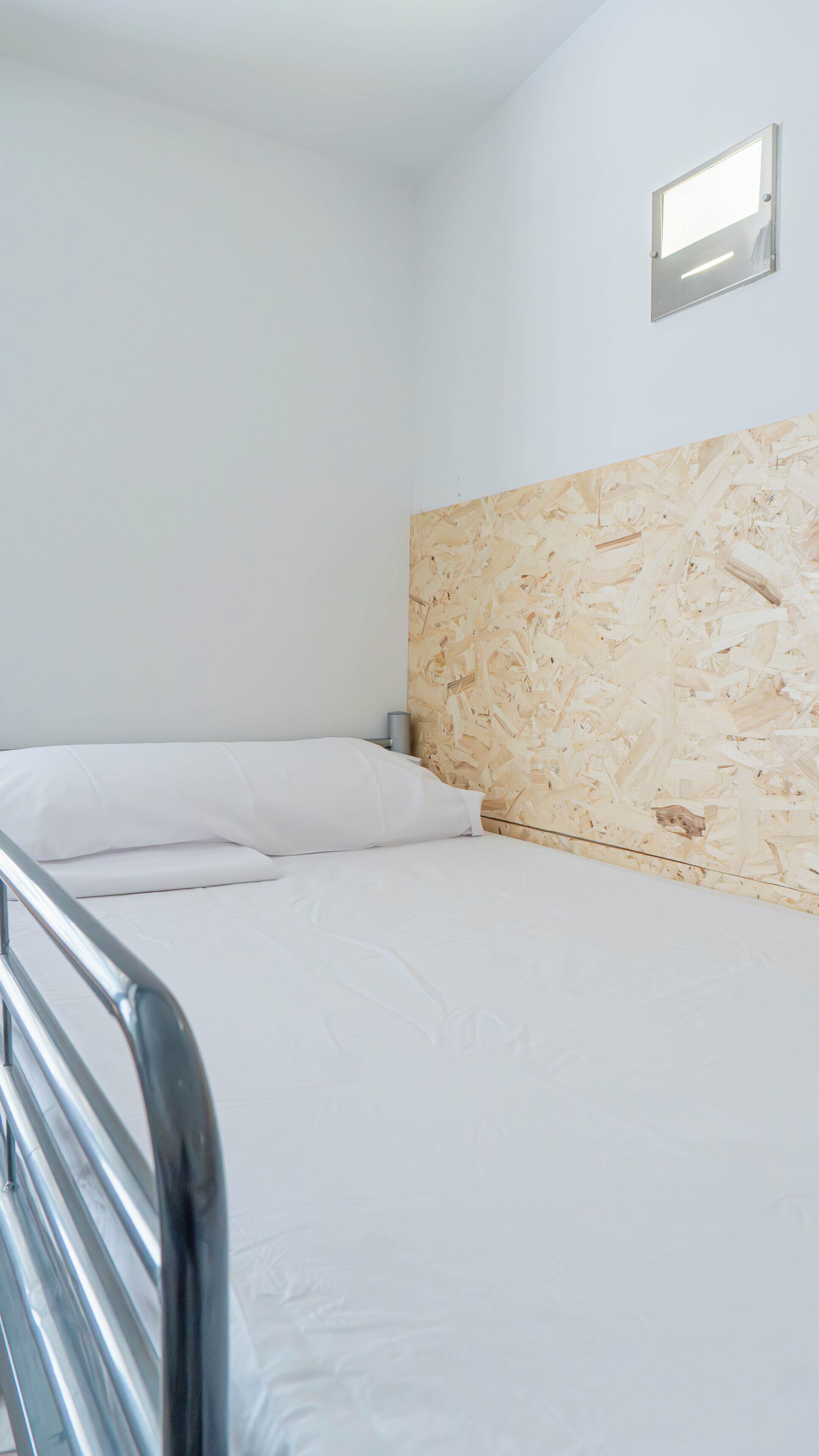 Een eenpersoonsbed met wit beddengoed in een kamer met een houten hoofdbord en een klein raam.