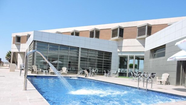 Una piscina con fuente, rodeada de sillas de descanso y un edificio moderno con grandes ventanales.
