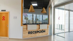 Zona de recepción con mostrador de madera, un cartel que dice “RECEPCIÓN” y una puerta de cristal que da al exterior.