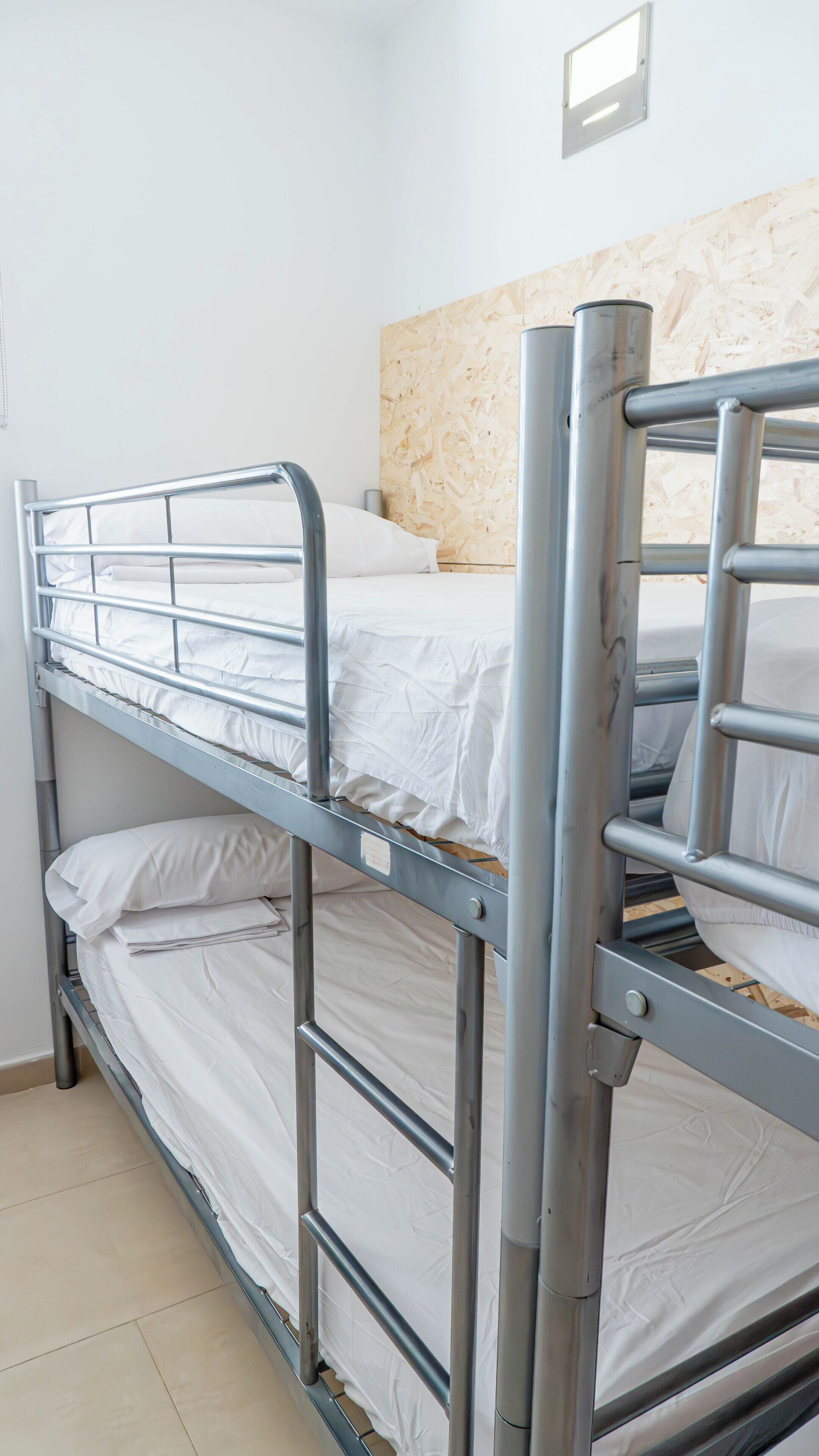 A bunk bed with metal railings and white bedding in a room with a wooden wall and tiled floor.