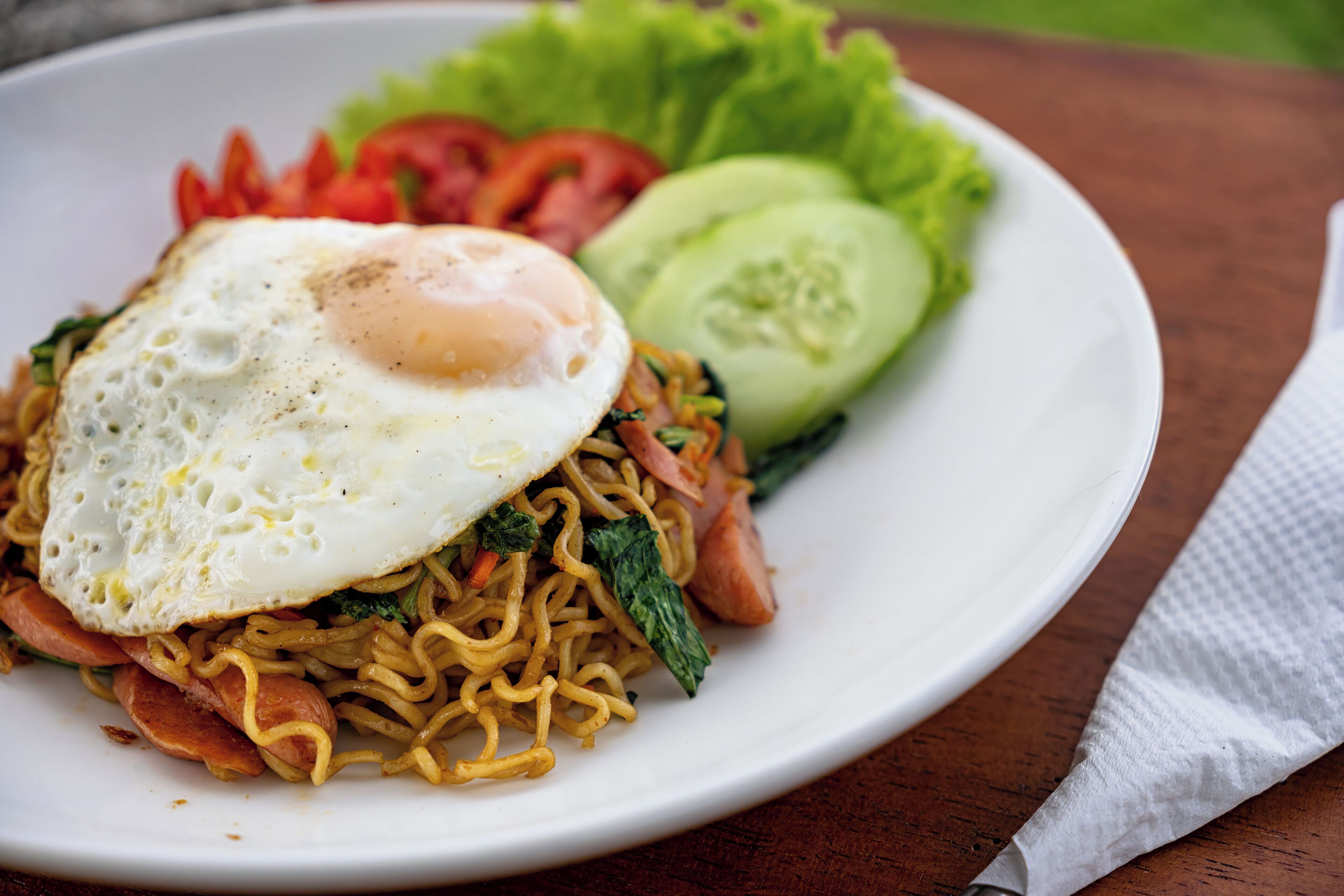 A plate of noodles topped with a fried egg, accompanied by sliced tomatoes, cucumbers, and lettuce.
