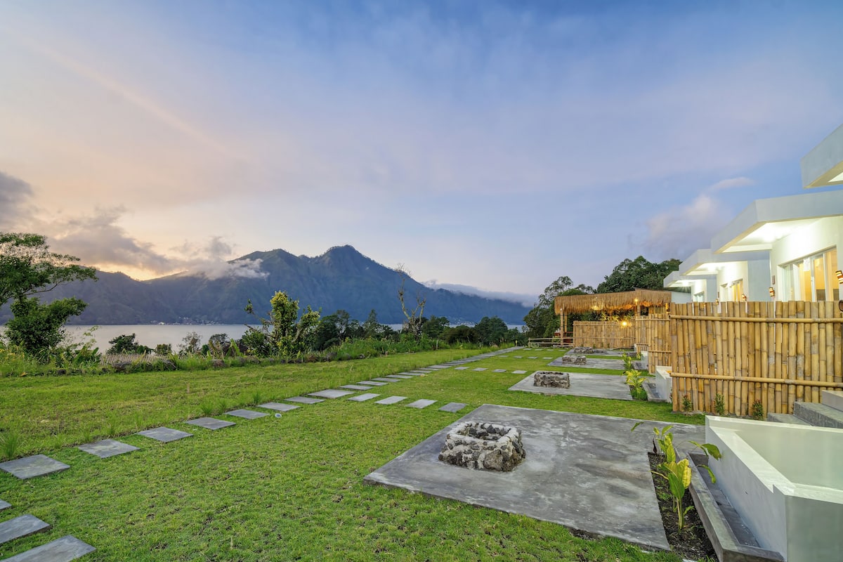 A modern hotel with a stone pathway leading to the entrance, surrounded by lush greenery and a view of mountains and a lake.