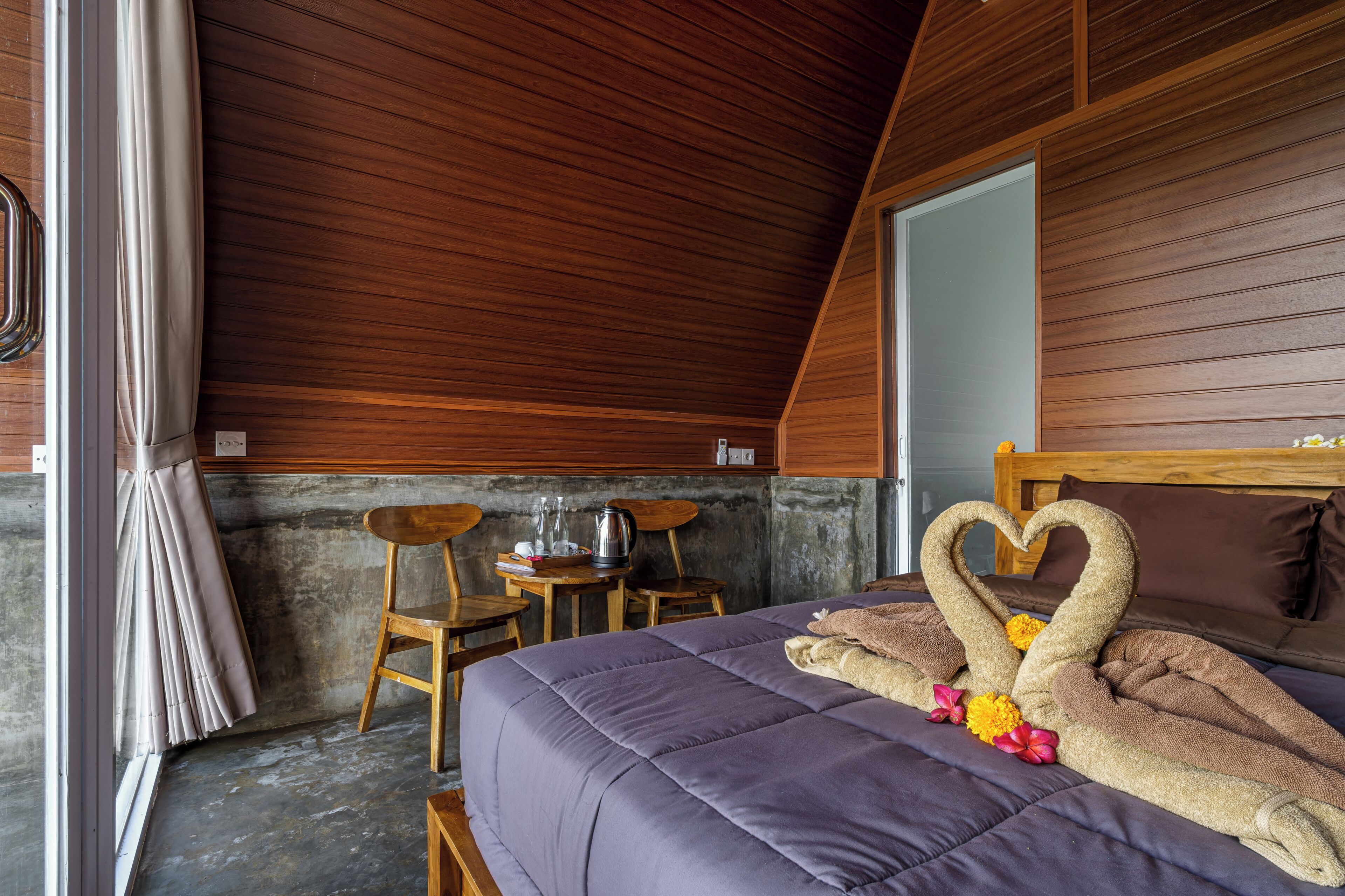 A room with a bed, wooden ceiling, a small table with chairs, and a window with curtains.