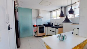 A modern kitchen with white cabinets, a stove, and a dining area with a white table and yellow chairs. - COBAR0110 - Edifício Beta (Salvador)