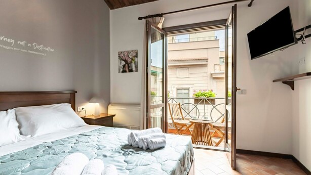 Une chambre d’hôtel comprenant un lit, une télévision, un balcon avec une table et des chaises, et un mur orné d’une citation.