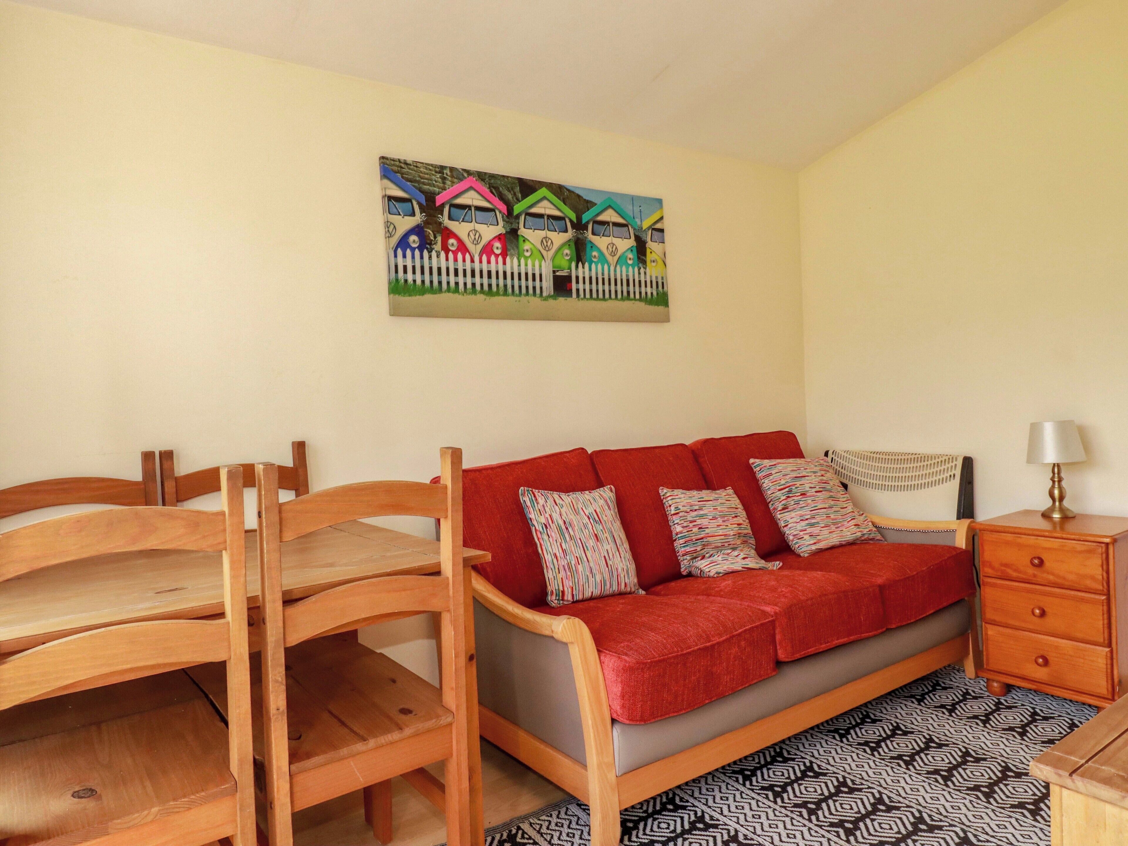 A room with a red sofa, wooden chairs, a patterned rug, a nightstand with a lamp, and a painting of colorful houses on the wall.