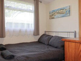 Un dormitorio con cama, ventana con cortinas, un cuadro en la pared y una mesita de noche de madera.