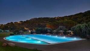Une grande piscine extérieure, dotée de nombreux chaises longues et parasols, entourée d’un jardin aménagé et bordée d’une colline en arrière-plan.