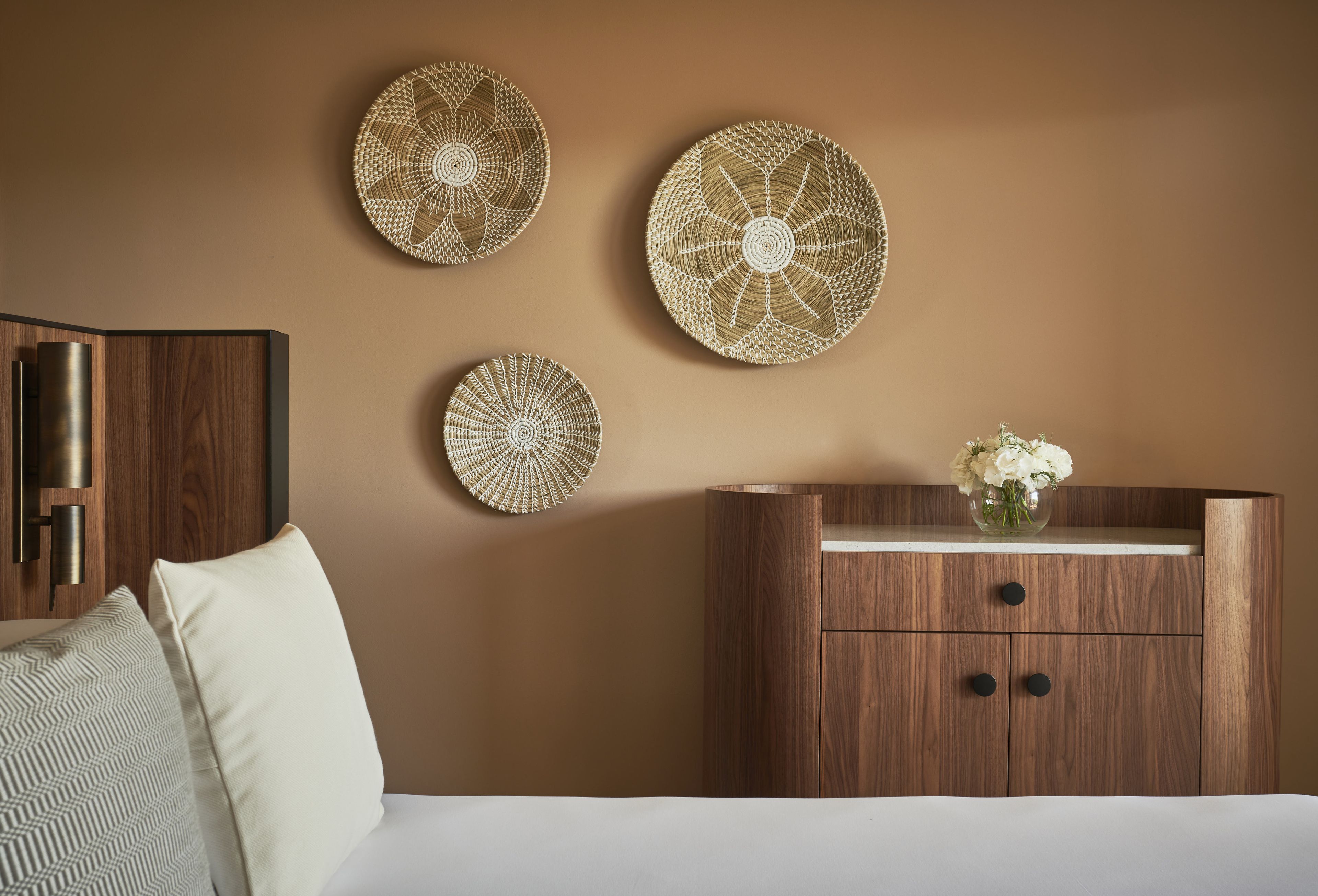 Un dormitorio con un aparador de madera, una cama con ropa blanca y tres piezas de decoración mural tejida.