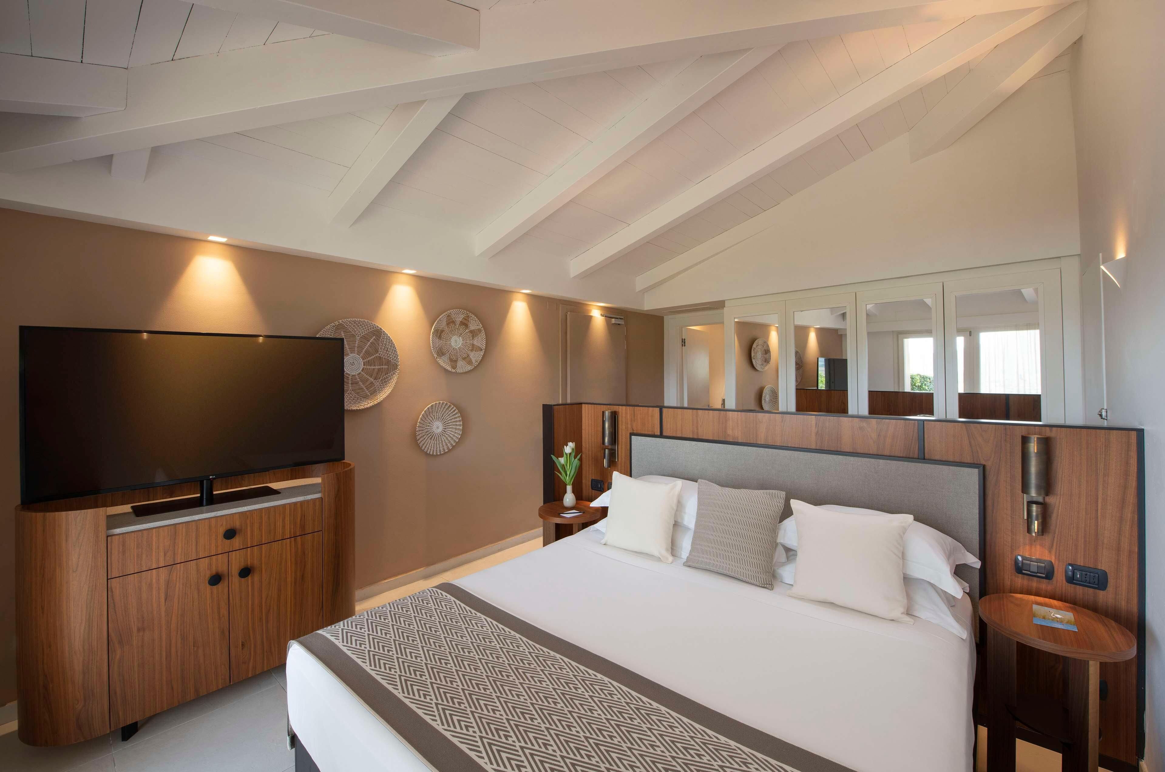 A modern hotel room with a large bed, a flat-screen TV, wooden furniture, and a ceiling with exposed beams.