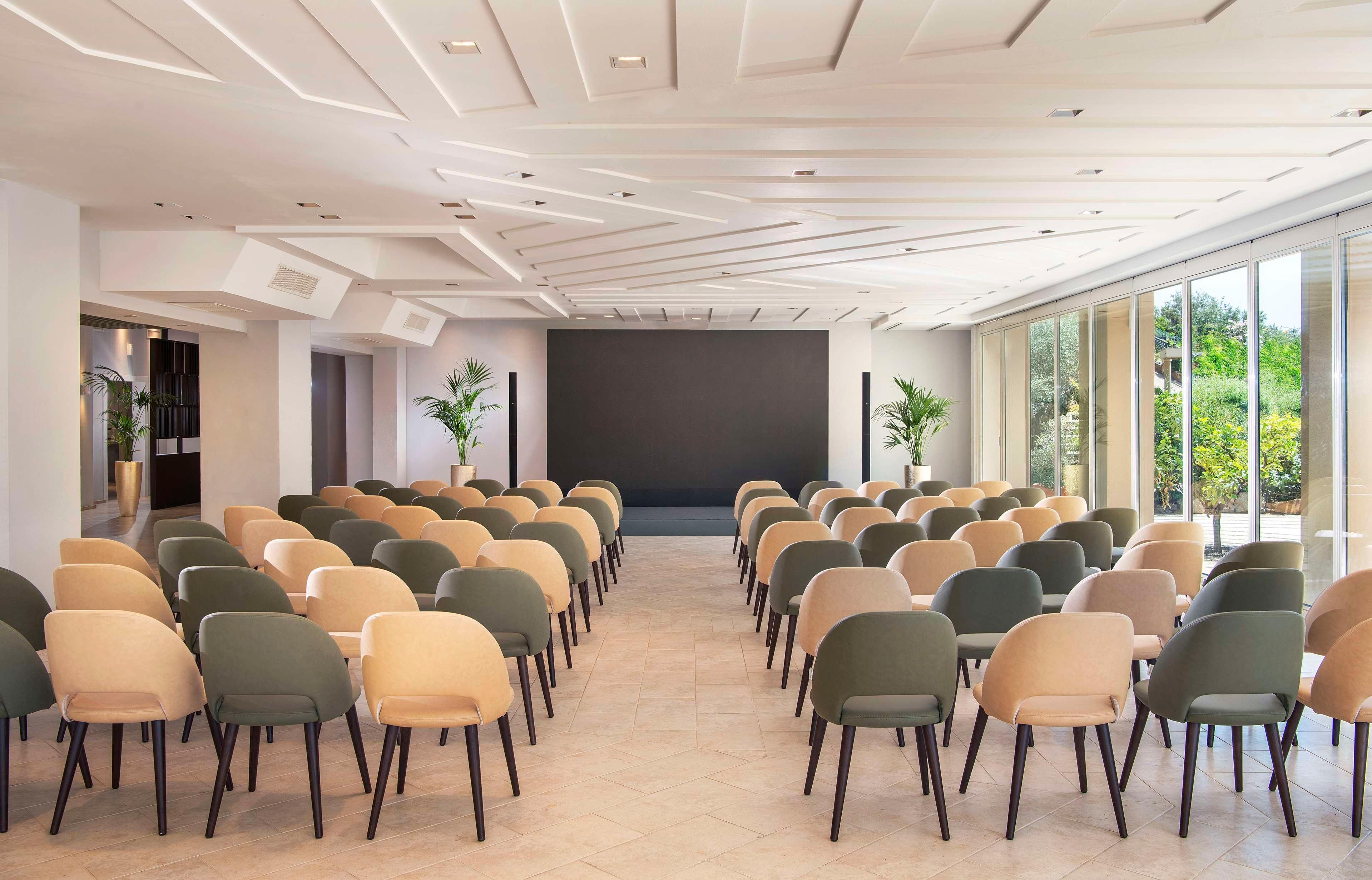 Ein Konferenzraum mit Reihen von Stühlen, einem großen Bildschirm und großen Fenstern mit Blick auf Grünflächen.